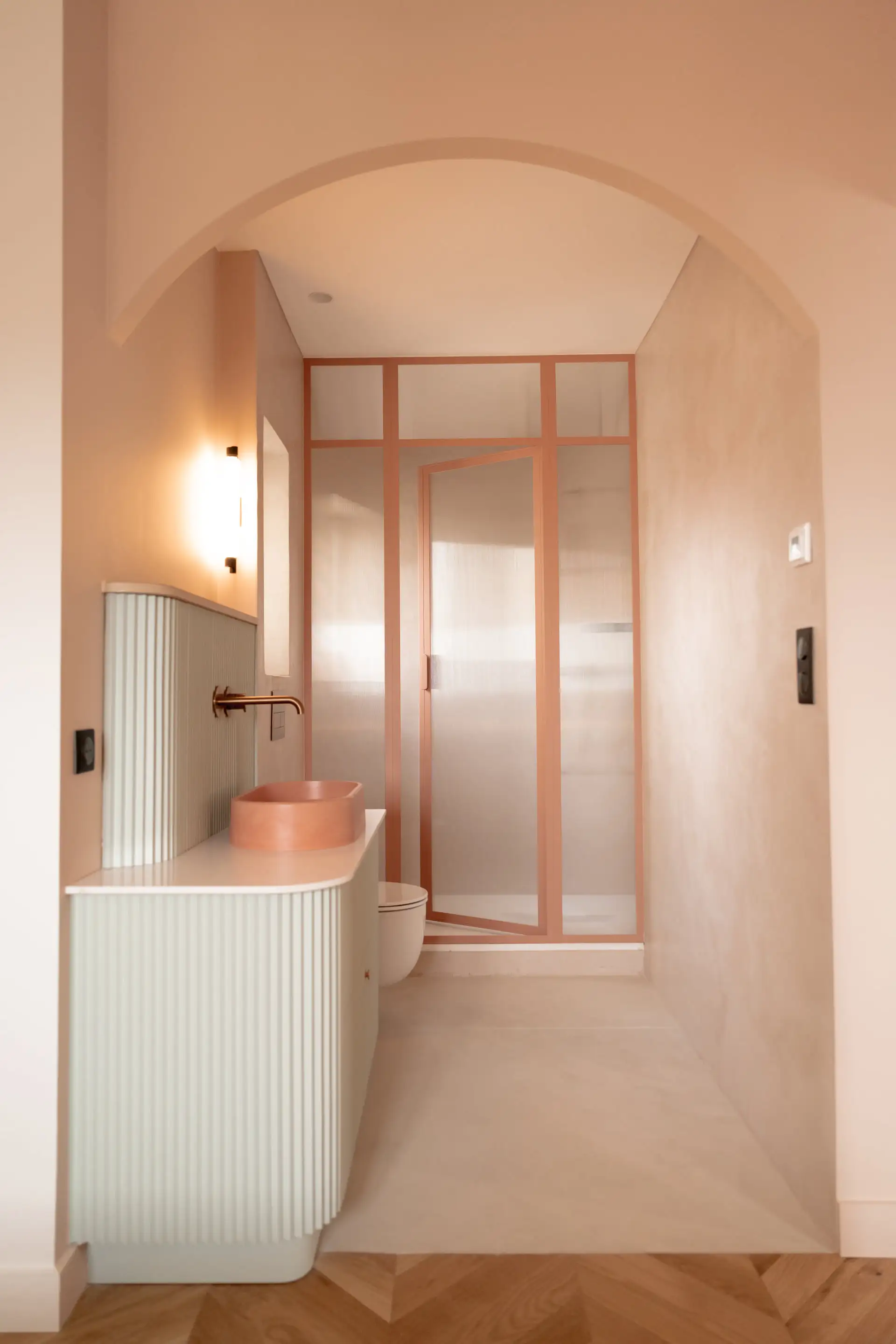 Salle d’eau rose poudré sous une arche, verrière et robinetterie cuivrée
