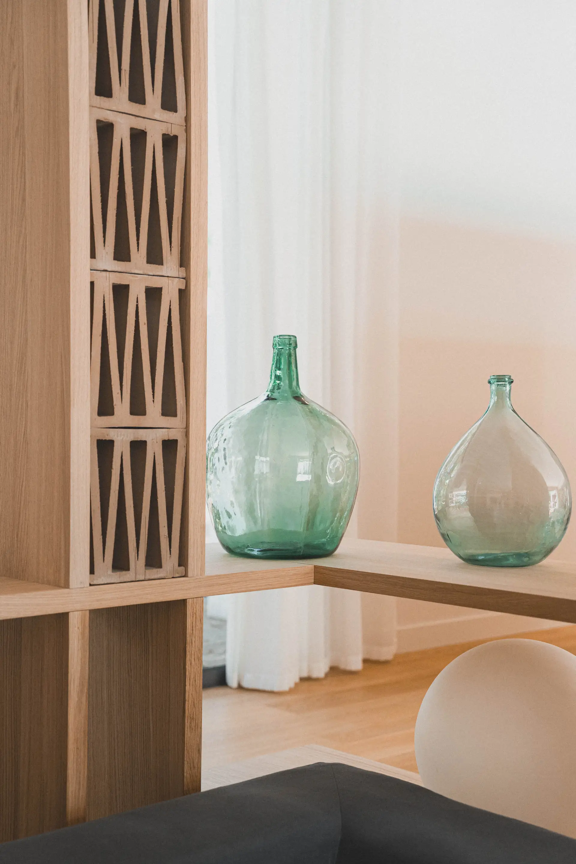 Claustra en bois ajouré traversée par la lumière, dames-jeannes en verre vert sur une étagère